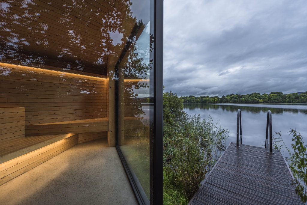 Interior of a modern wooden sauna with ambient lighting, overlooking a serene lake and greenery outside.
