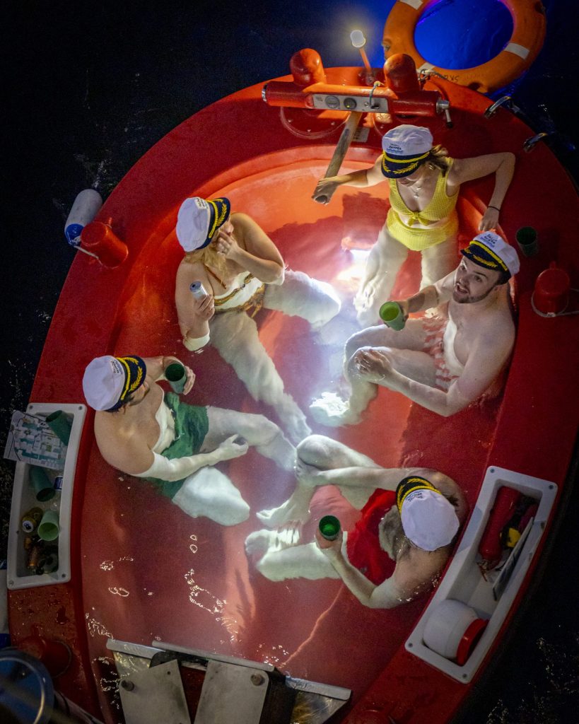 A group of people relaxing in a red hot tub-style boat, wearing nautical hats, enjoying drinks and having fun.