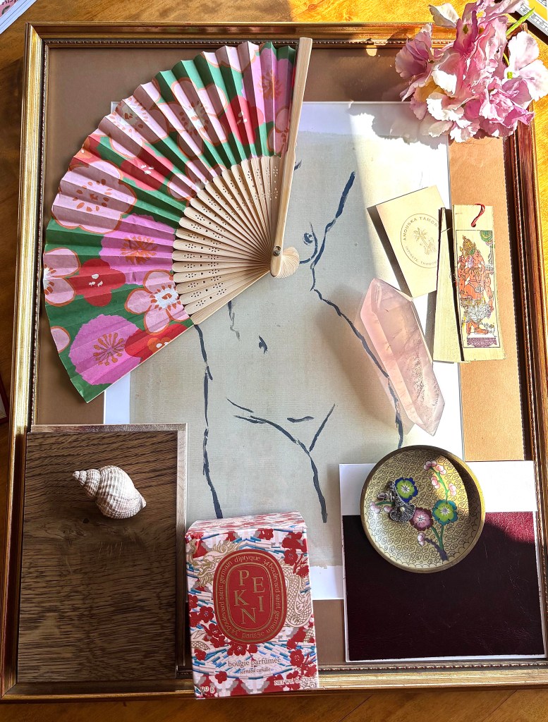 A stylish flat lay featuring a colorful fan, a shell, a pink crystal, and a decorative plate, arranged on a wooden surface with an art print and various decorative items.