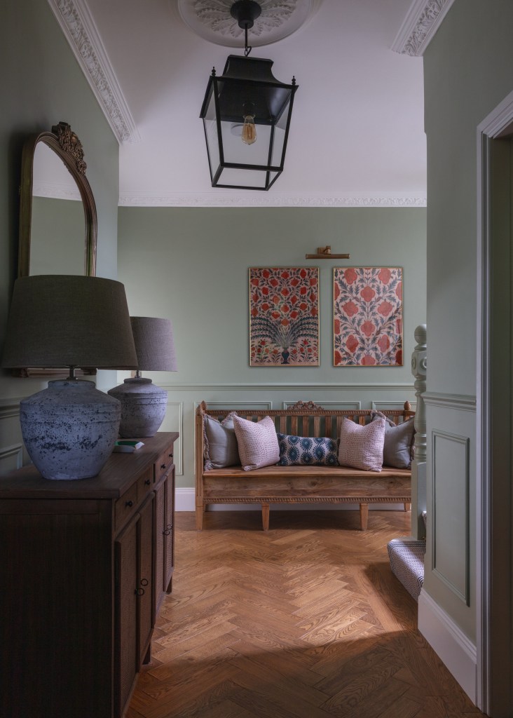 A warmly decorated hallway featuring a combination of earthy tones, a wooden bench with decorative pillows, and elegant wall art. The space is illuminated by a stylish lantern and showcases herringbone flooring.
