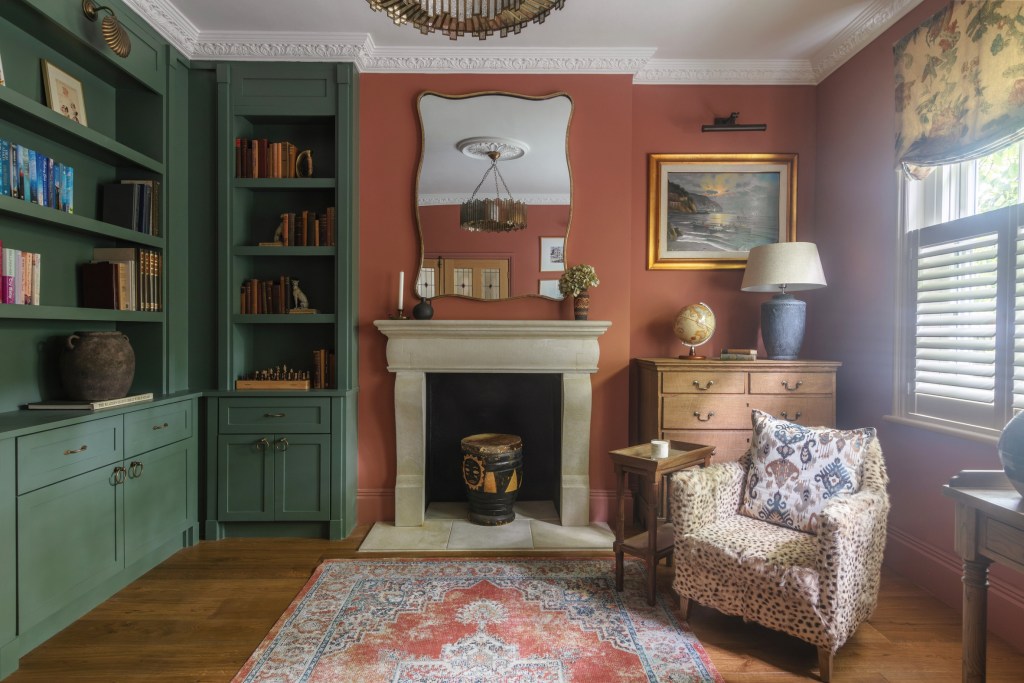 A cozy living room featuring green bookshelves, a terracotta wall, and a stone fireplace. Furnished with a leopard-print chair, a wooden side table, and decorative items like a globe and artwork.