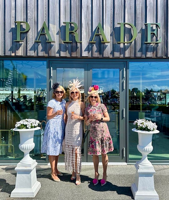 Three women in stylish outfits and hats stand in front of a venue sign labeled 'PARADE,' holding drinks with flowers in white planters beside them.