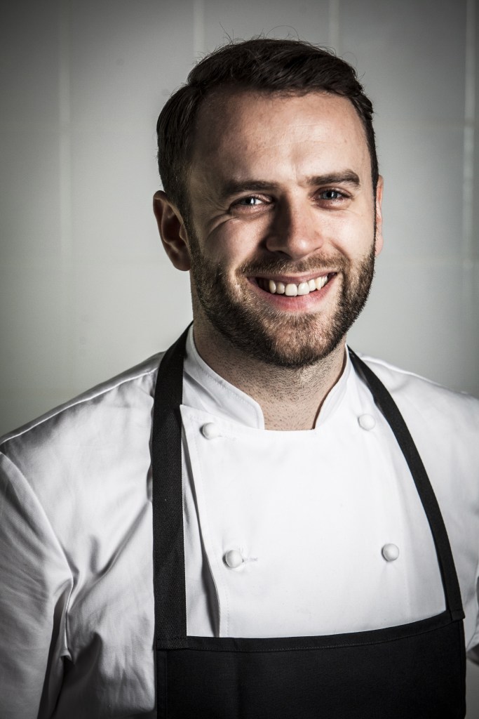 A smiling chef wearing a white uniform and a black apron, standing in a kitchen setting.