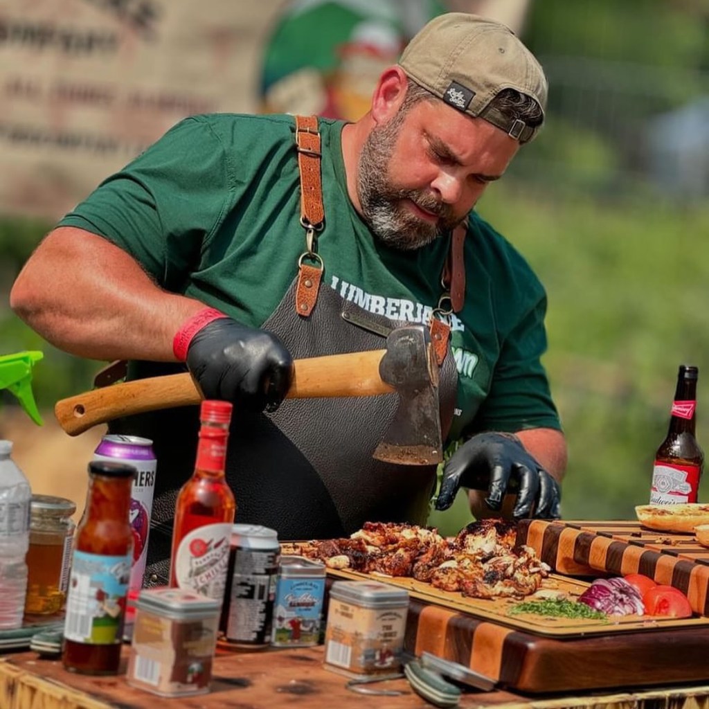 A chef in a green shirt and apron using a large axe to prepare food at an outdoor setting, with various beverages and ingredients displayed on a wooden table.