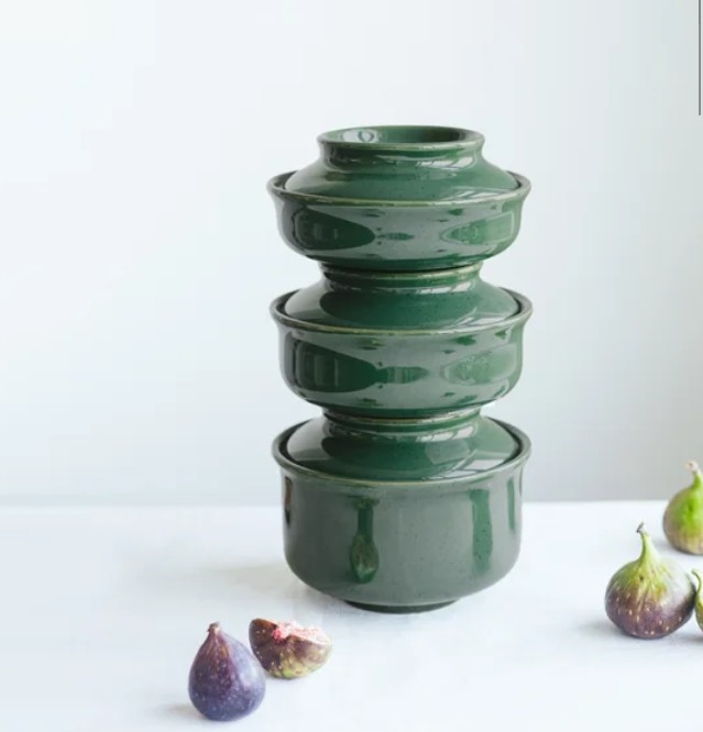 Three stackable green stoneware bowls arranged vertically on a white surface, alongside fresh figs.