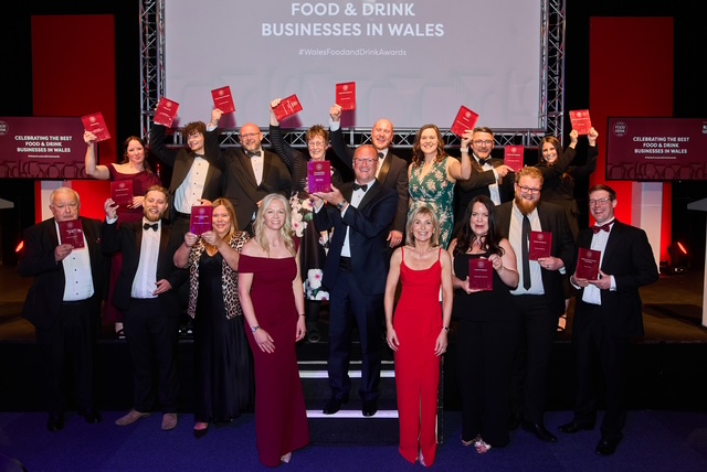A group of award winners and presenters celebrating at the Wales Food and Drink Awards, holding red award plaques, with a festive backdrop.