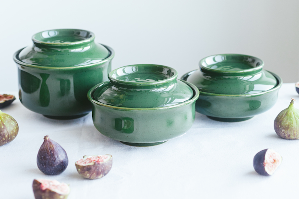 Three stacked green stoneware bowls with lids, surrounded by figs on a light background.