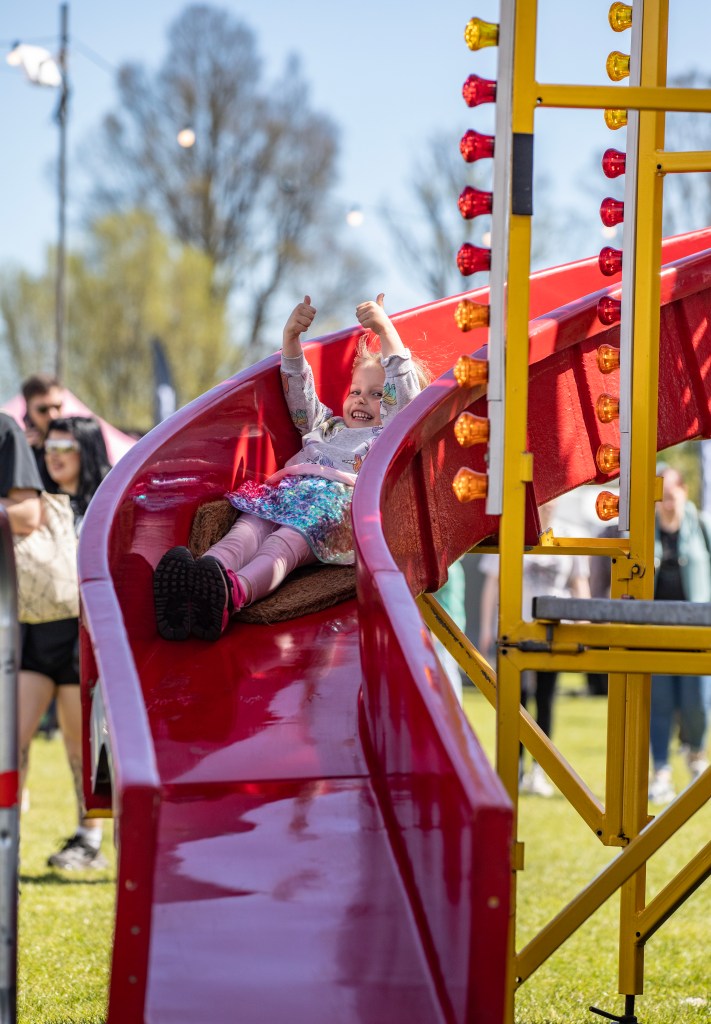 A young child joyfully sliding down a red playground slide, giving a thumbs-up while enjoying the ride, with people in the background.