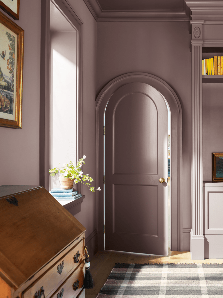 An elegantly styled interior featuring a soft purple wall, an arched door, artwork, and a wooden desk, with a potted plant on a window sill.