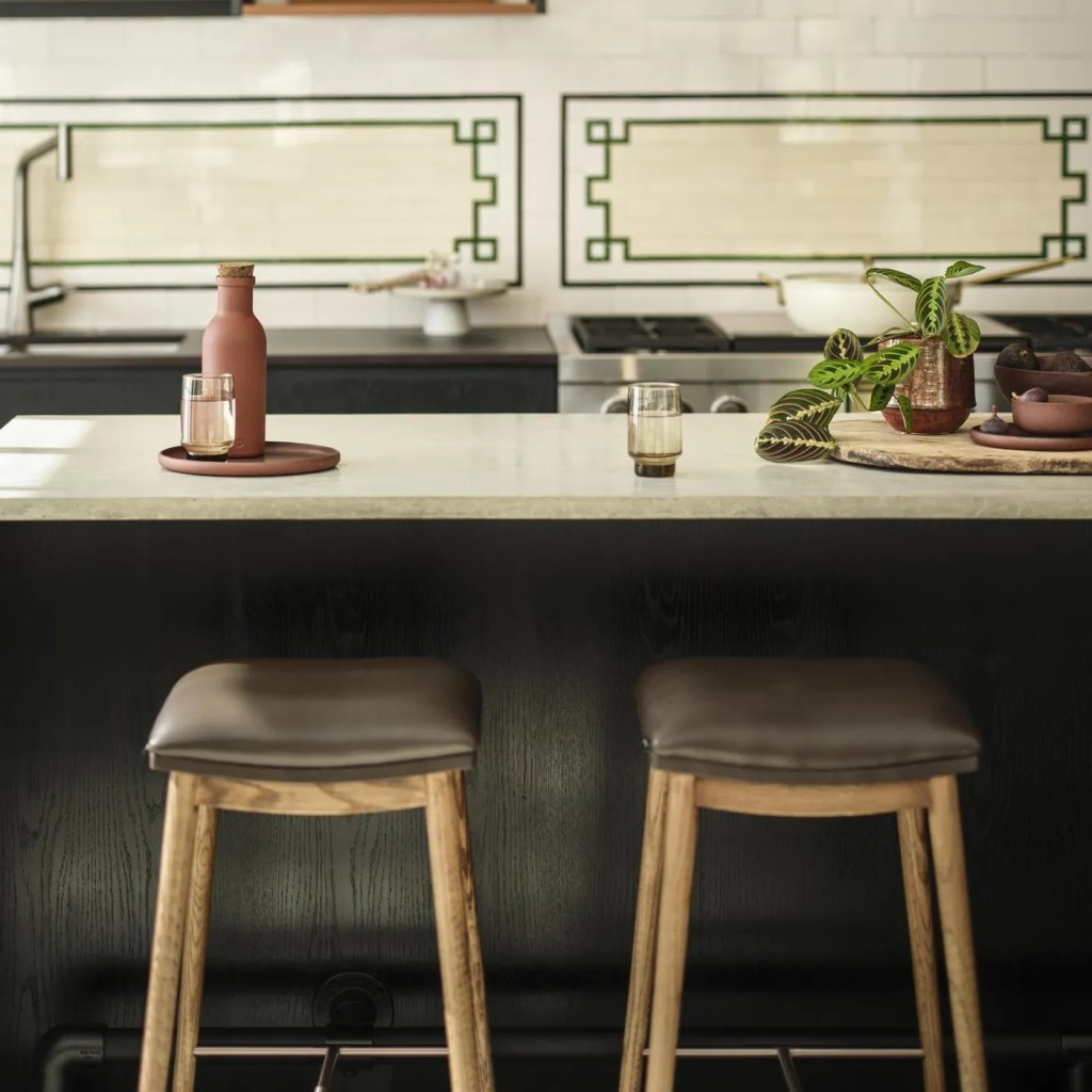 A modern kitchen island featuring two wooden stools with brown cushions, a ceramic vase with a plant, a glass of beverage, and a terracotta bottle, set against a stylish patterned backsplash.