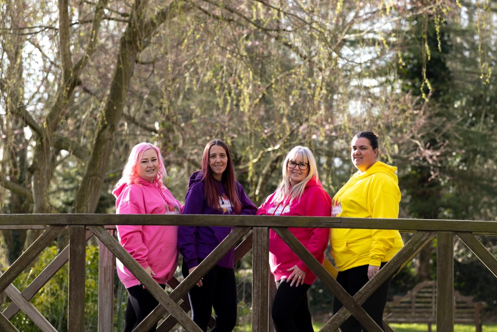 A group of four women wearing colorful hoodies, standing on a wooden bridge with lush greenery and trees in the background.