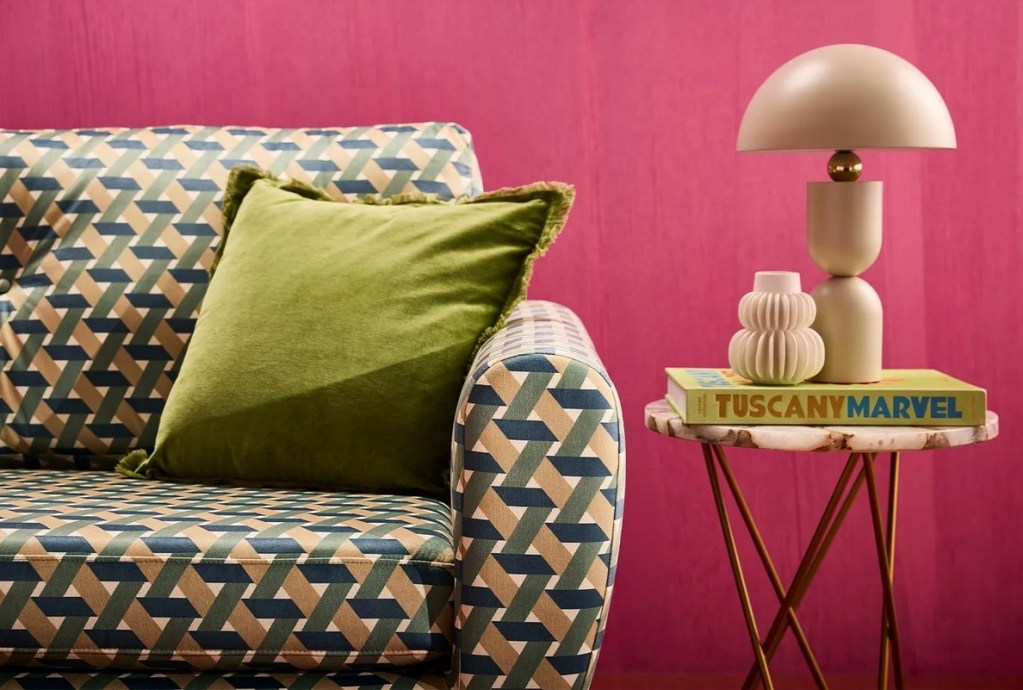A colorful sofa with a geometric print in blue, beige, and green, adorned with a green velvet pillow, next to a small round table featuring a white lamp and books.
