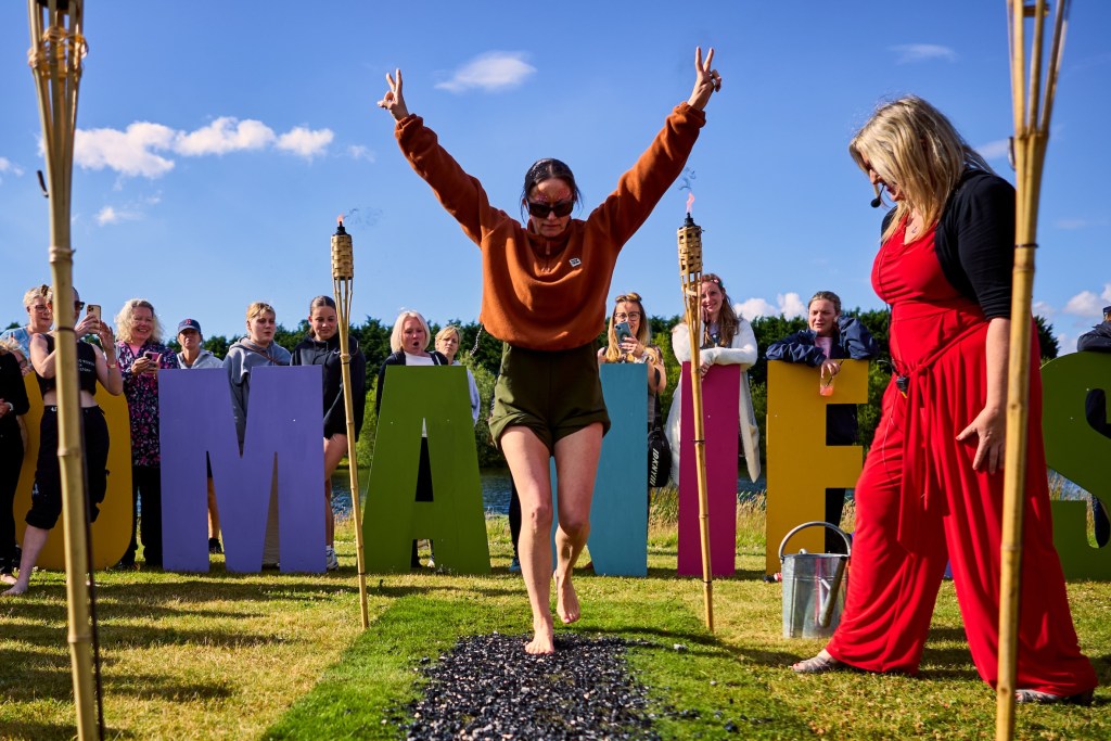 A woman joyfully crossing a firewalk event at the WOMANIFEST festival, with vibrant letters spelling 'WOMANIFEST' in the background, surrounded by an engaged audience.