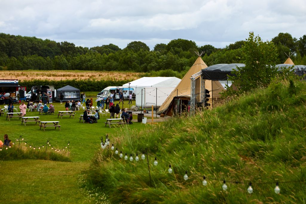 A vibrant outdoor festival scene featuring tents, food stalls, and attendees enjoying the activities in a grassy field.