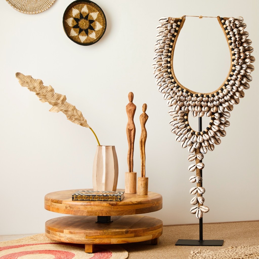 A wooden coffee table with a decorative magazine, a stylized vase with a dried plant, two carved wooden figures, and a large shell ornament displayed on a stand against a neutral wall.