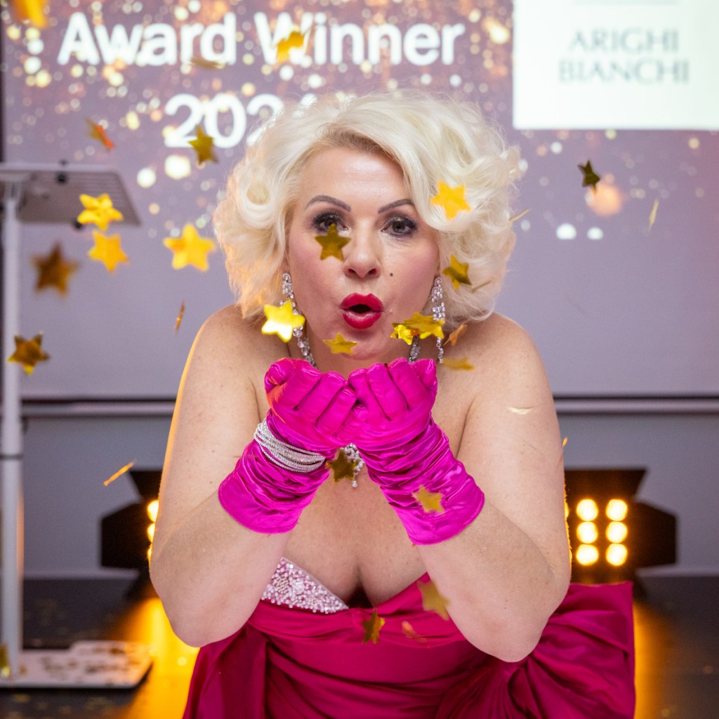 A glamorous individual celebrating an award win, blowing gold star confetti with a joyful expression, wearing a sparkling pink gown and matching gloves.