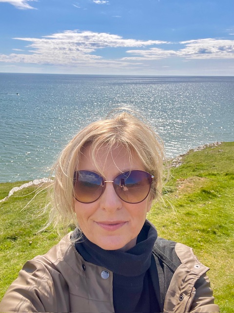 A person with sunglasses standing on a grassy shoreline overlooking a calm sea under a cloudy blue sky.
