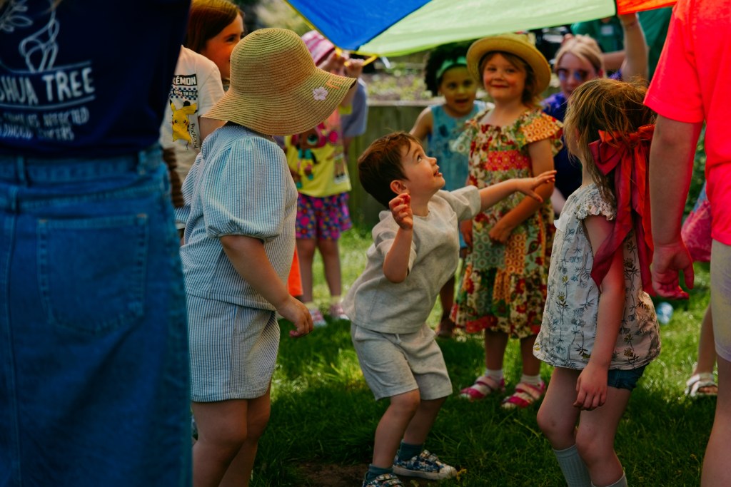 A group of children playing joyfully under a colorful parachute on a sunny day, showcasing smiles and playful interaction.