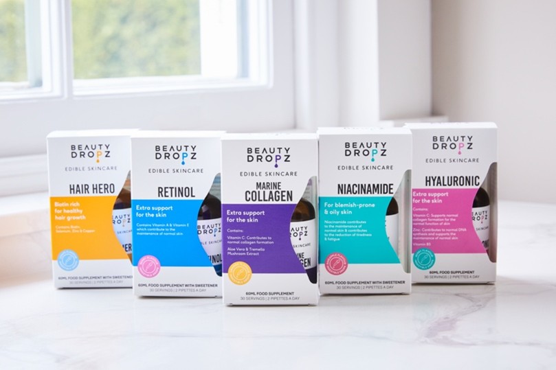 A display of five Beauty Dropz edible skincare supplements, including Marine Collagen, Niacinamide, Hair Hero, Retinol, and Hyaluronic drops, arranged neatly on a countertop with a window in the background.