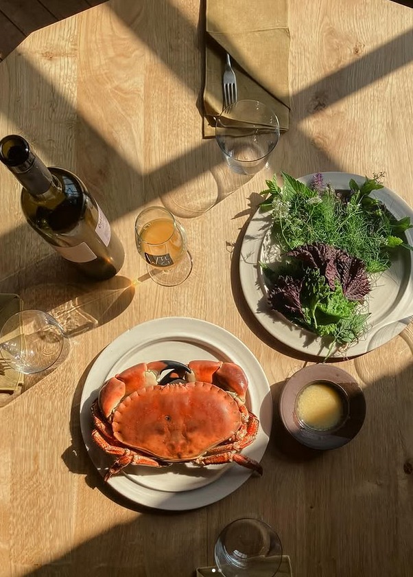 A wooden table setting featuring a cooked crab on a white plate, a side salad with herbs and greens on a beige plate, a bottle of wine, glasses, and a round brown bowl.