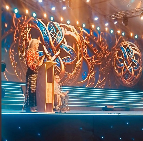 A woman speaking at a podium on stage during a cultural festival, with a decorative backdrop featuring intricate designs and bright stage lighting.