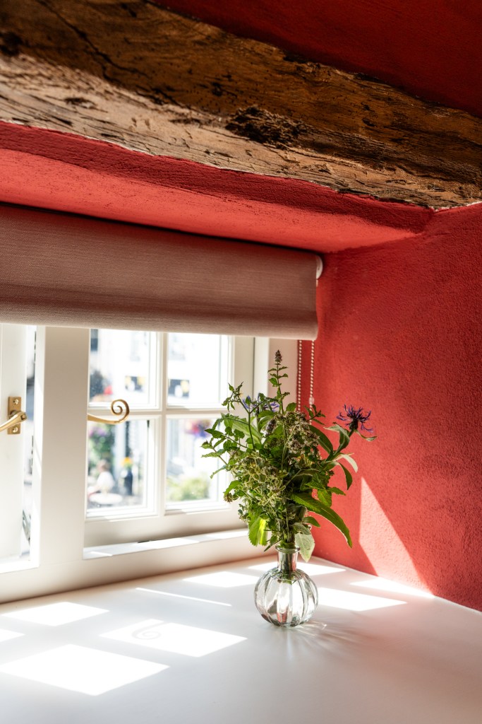 A vase with fresh flowers sits on a white windowsill against a colorful wall, featuring a wooden beam above and soft sunlight streaming through the window.