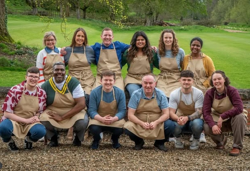 A group of ten bakers, wearing aprons, smiles together outdoors in a green setting, showcasing camaraderie and a shared passion for baking.