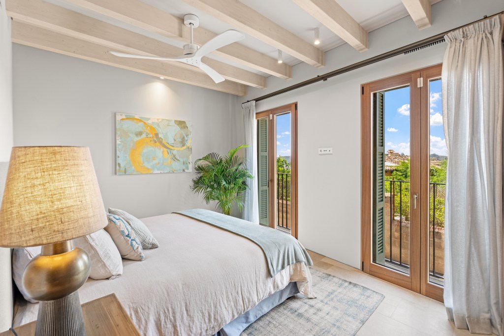 A cozy bedroom with wooden beams, a soft bed with neutral linens, a bedside lamp, and large windows showcasing greenery and a scenic view.