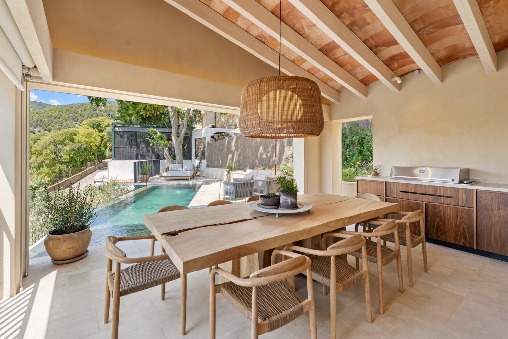 Modern dining area with a wooden table and chairs, opening to a view of an infinity pool and lush outdoor surroundings in the backdrop.