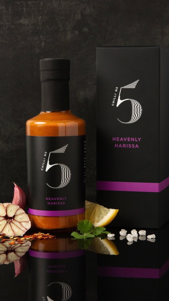 A bottle of Heavenly Harissa sauce with a sleek design, accompanied by a box and garnished with garlic, lemon, coriander, and salt on a reflective surface.