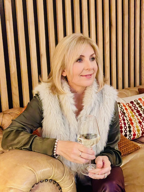 A person sitting on a leather couch, holding a glass of wine, wearing a stylish outfit with a fur vest. The background features wooden paneling and colorful patterned cushions.