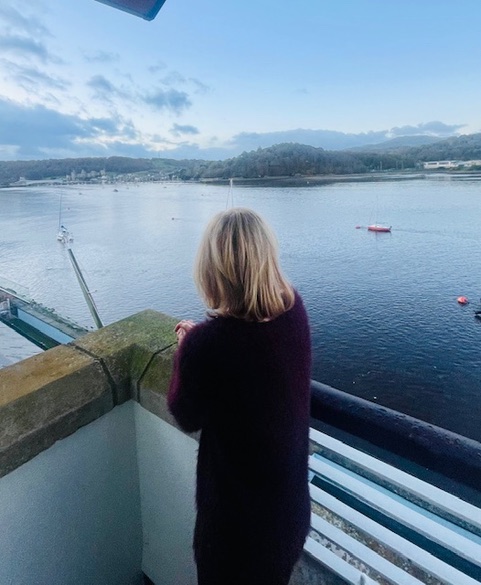A person standing on a balcony overlooking the calm waters of the Conwy Estuary, with boats visible in the distance and rolling hills in the background.