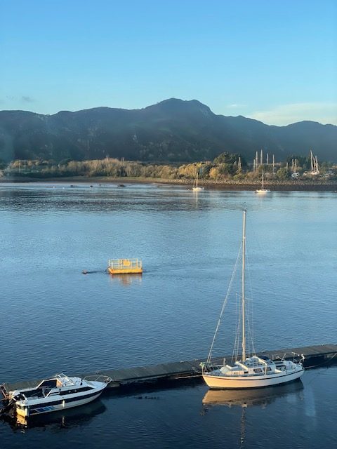 A serene view of the Conwy Estuary with two boats docked in the foreground, mountains in the background, and a clear blue sky.
