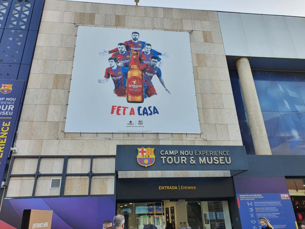Exterior view of the Camp Nou Experience entrance, featuring a large promotional banner with players in FC Barcelona jerseys and the text 'FET A CASA'.