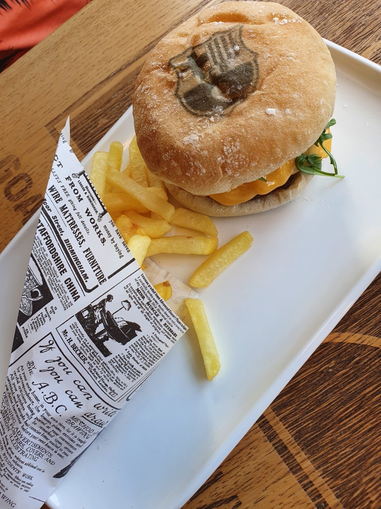 A plate featuring a sandwich with a stamped design on the bun, filled with cheese and greens, accompanied by a portion of French fries served in a printed paper cone.