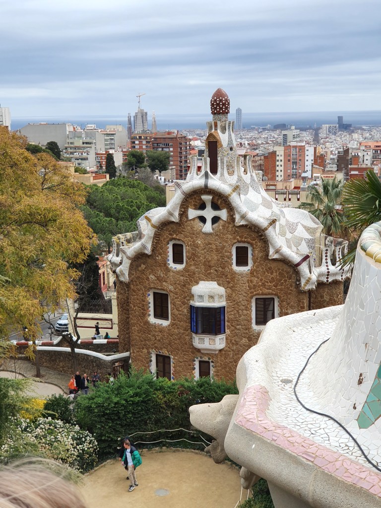A view of the colorful architectural design of Park Güell in Barcelona, featuring a unique gingerbread-like building with mosaic details, surrounded by greenery and the city skyline in the background.