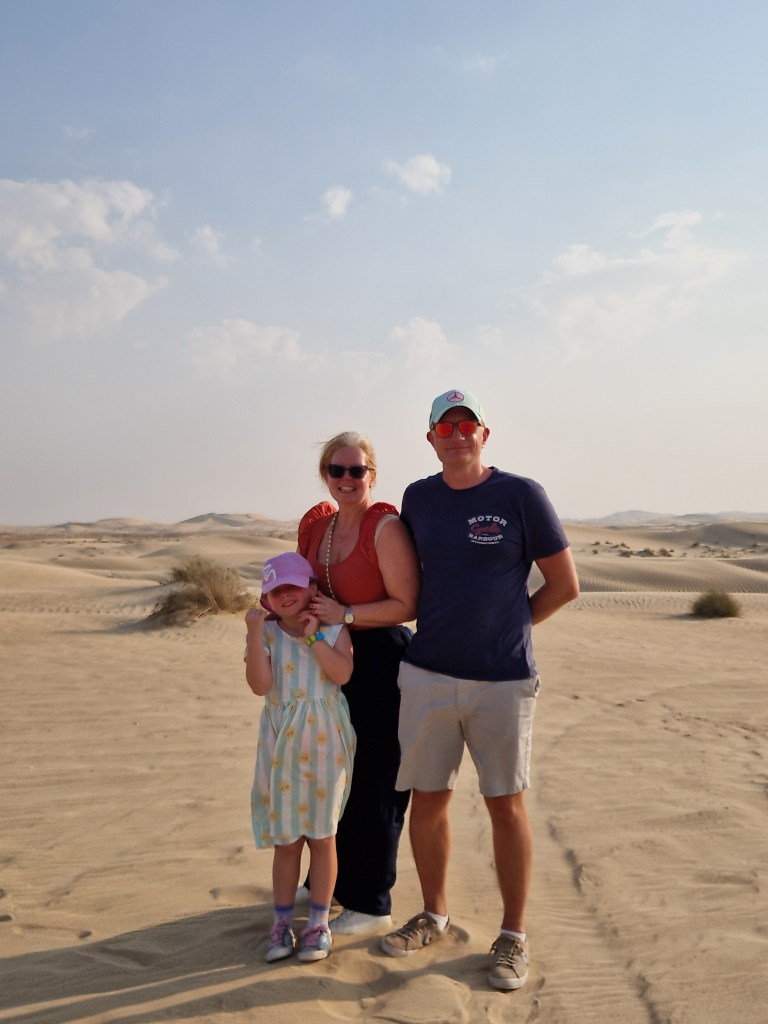 A family of three poses together in a sandy desert landscape, with light clouds in the sky.