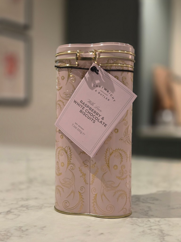 A decorative pink tin containing raspberry and white chocolate biscuits, with a tag displaying the product name and weight.