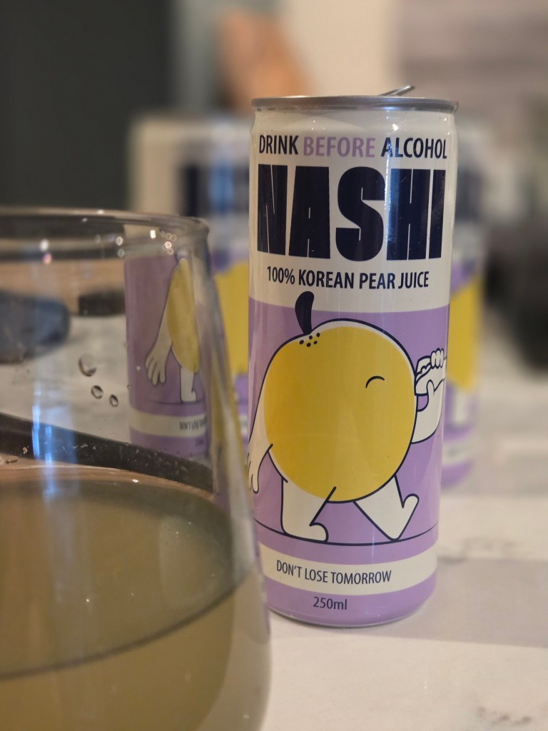 A can of Nashi 100% Korean pear juice, labeled 'Drink Before Alcohol,' with a cartoon character illustration. A glass partially filled with a light-colored liquid is in the foreground.