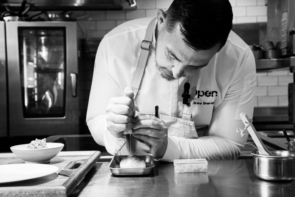 A chef meticulously plating a dish in a professional kitchen, using a small tool to refine presentation.