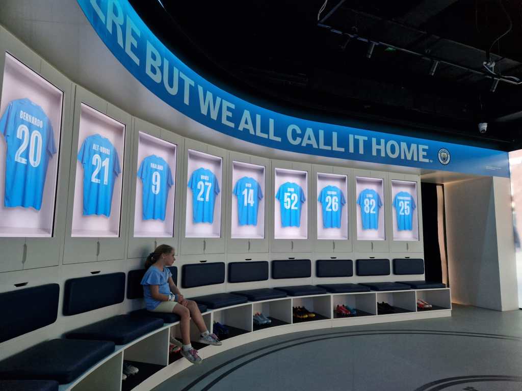 A modern locker room display featuring Manchester City football jerseys with player names and numbers, along with a child sitting on a bench.