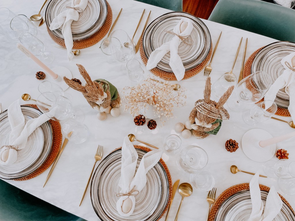 A beautifully arranged dining table featuring woven placemats, elegant tableware, and decorative bunny-themed napkin holders. The table is adorned with glasses, golden cutlery, dried flowers, and pine cones, set against a white marble surface.