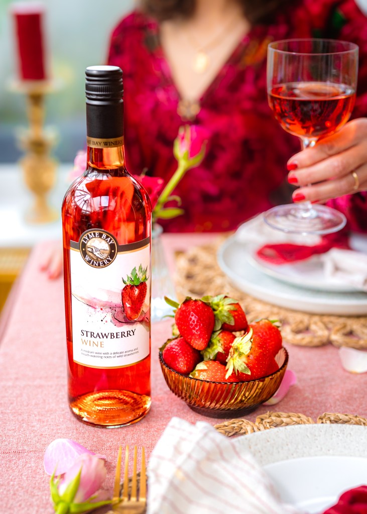 A bottle of strawberry wine sits on a pink tablecloth accompanied by a bowl of fresh strawberries. A person in the background holds a glass of the wine, with a candle and decorative elements enhancing the setting.