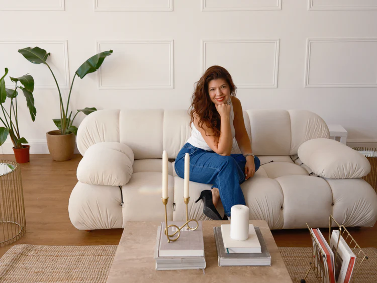 A woman seated on a cream-colored sofa, wearing a white top and blue jeans, smiling while resting her chin on her hand. A coffee table in front of her features a white candle and decorative items. Green plants are visible in the background.