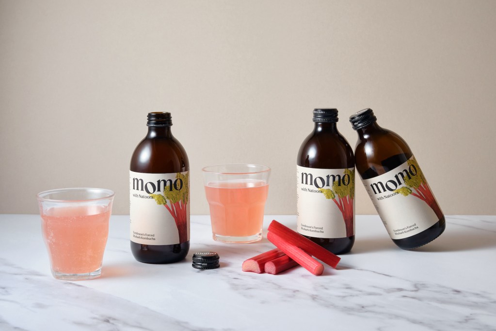 Bottles of Momo kombucha next to two glasses of pink liquid on a marble surface, with fresh rhubarb pieces in the foreground.