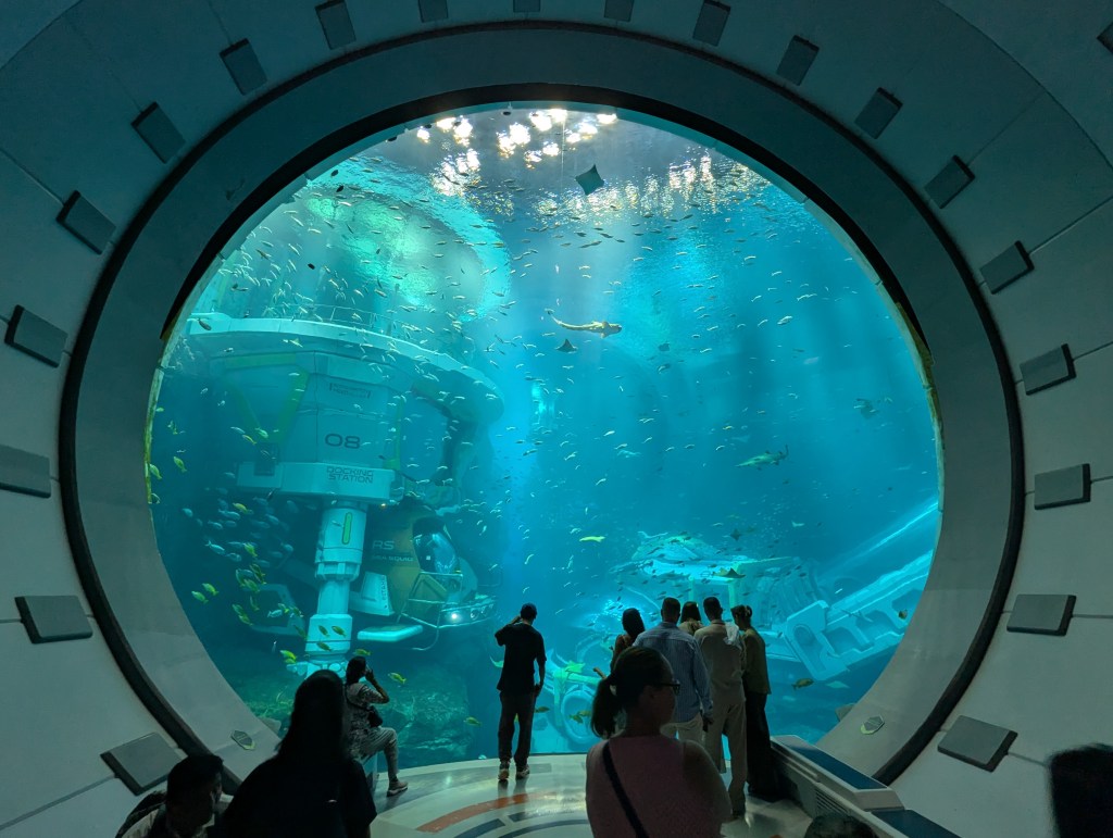 View of an underwater aquarium through a circular window, showcasing various colorful fish swimming around a submerged station.