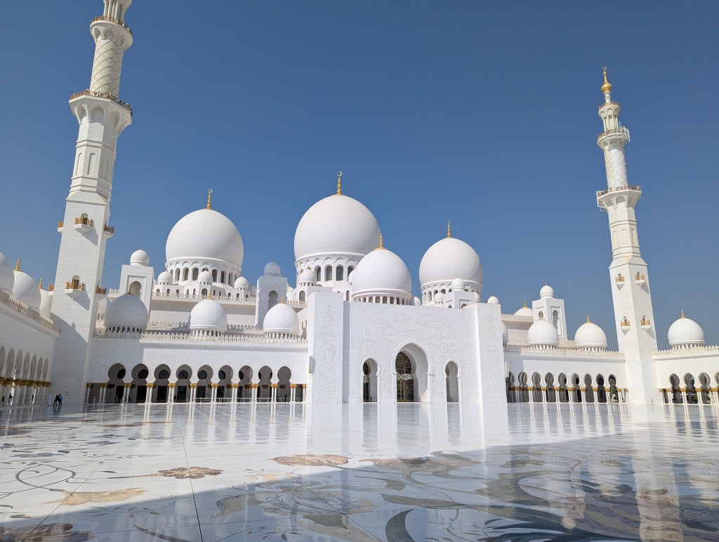 A wide view of the Sheikh Zayed Grand Mosque, showcasing its stunning white architecture, large domes, and a tall minaret against a blue sky.