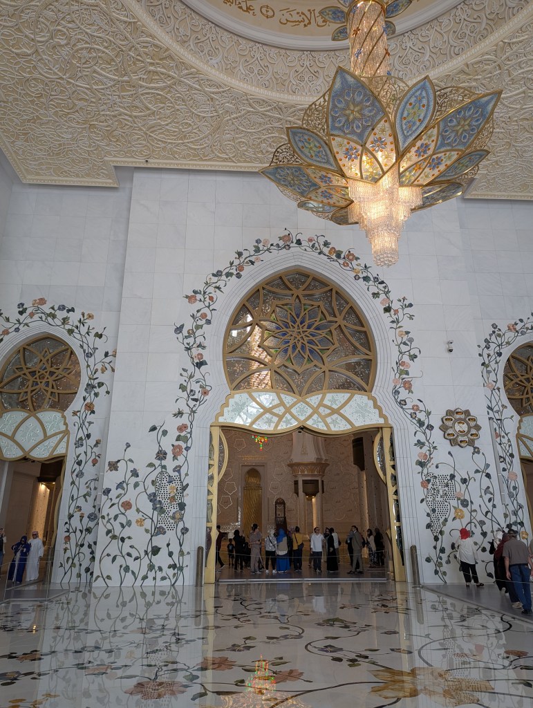 Interior view of a lavishly decorated mosque showcasing intricate floral patterns on the walls and a grand chandelier resembling a flower.