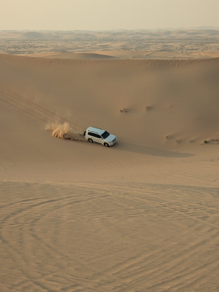 A white SUV driving through sandy dunes in a desert landscape, leaving a trail of dust behind.