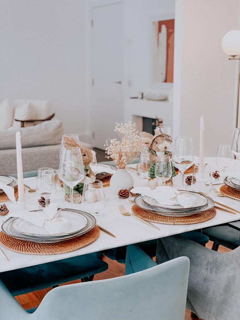 Elegant dining table set with decorative plates, glassware, and candles, featuring a floral centerpiece and pinecones, in a modern and stylish setting.